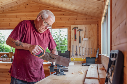 Superior Shed Workshop Man sawing a piece of timber at a workbench inside a spacious, timber-lined garden workshop.